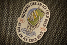 A die-cut sticker featuring a hand holding an ice-cold beer can above water, with the text 'NOTHING LIKE AN ICE COLD BEER AFTER AN ICE COLD BEER' around the edge, placed on a woven dark surface.