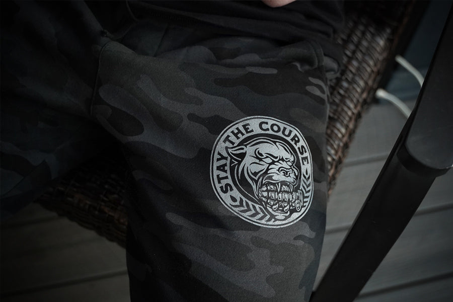 Black camouflage pants with a logo on the leg, featuring text and a graphic design.