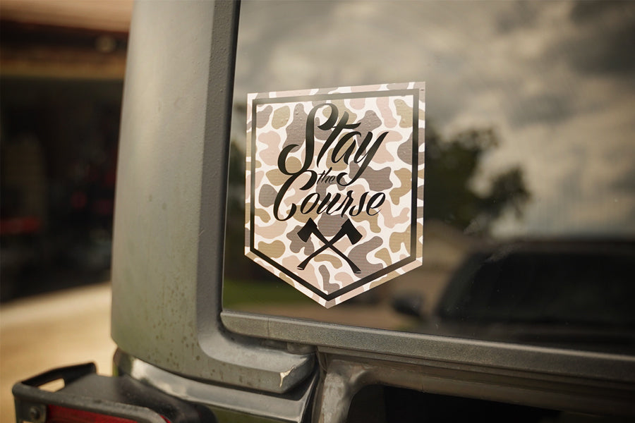 Camouflage-style 'Stay the Course' decal on a vehicle window with a blurred outdoor background.