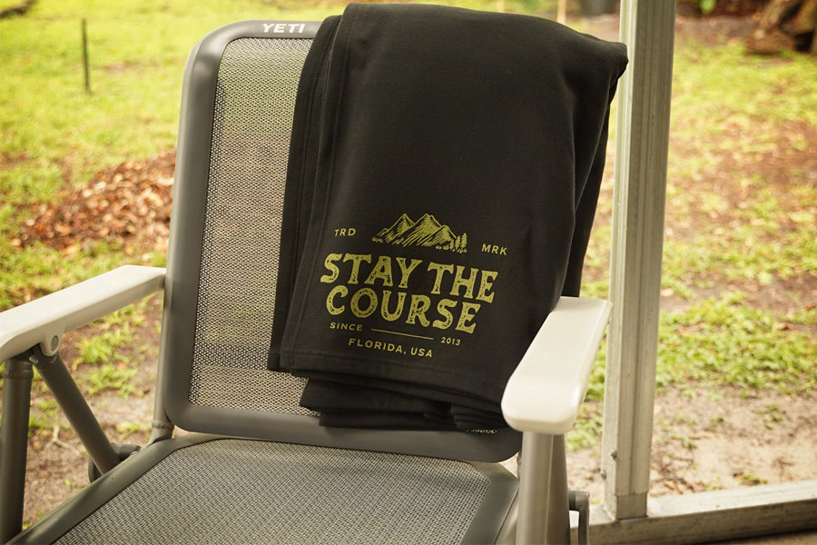 A black fleece blanket draped over a gray patio chair, featuring a green graphic that reads 'STAY THE COURSE' with mountains and 'FLORIDA, USA' on a porch with a grassy yard in the background.