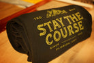 A rolled black blanket with distressed green text reading 'STAY THE COURSE', photographed on a wooden surface.