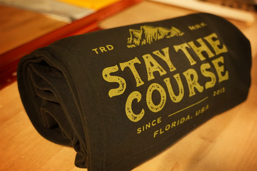 A rolled black blanket with distressed green text reading 'STAY THE COURSE', photographed on a wooden surface.