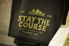 A black fleece blanket draped over a gray patio chair, featuring a green graphic that reads 'STAY THE COURSE' with mountains and 'FLORIDA, USA' on a porch with a grassy yard in the background.