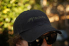 Black cap with 'Stay the Course' text worn by a person with sunglasses, blurred natural background