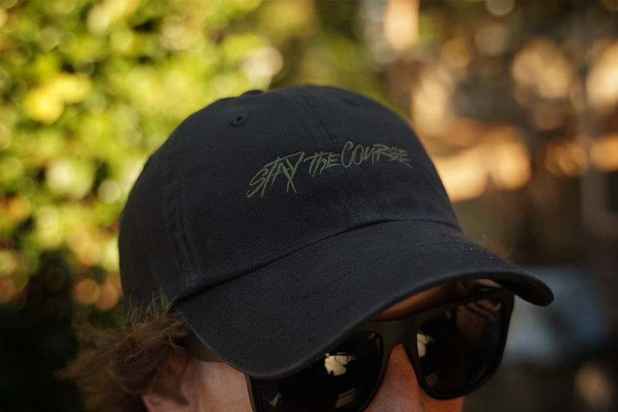 Black cap with 'Stay the Course' text worn by a person with sunglasses, blurred natural background