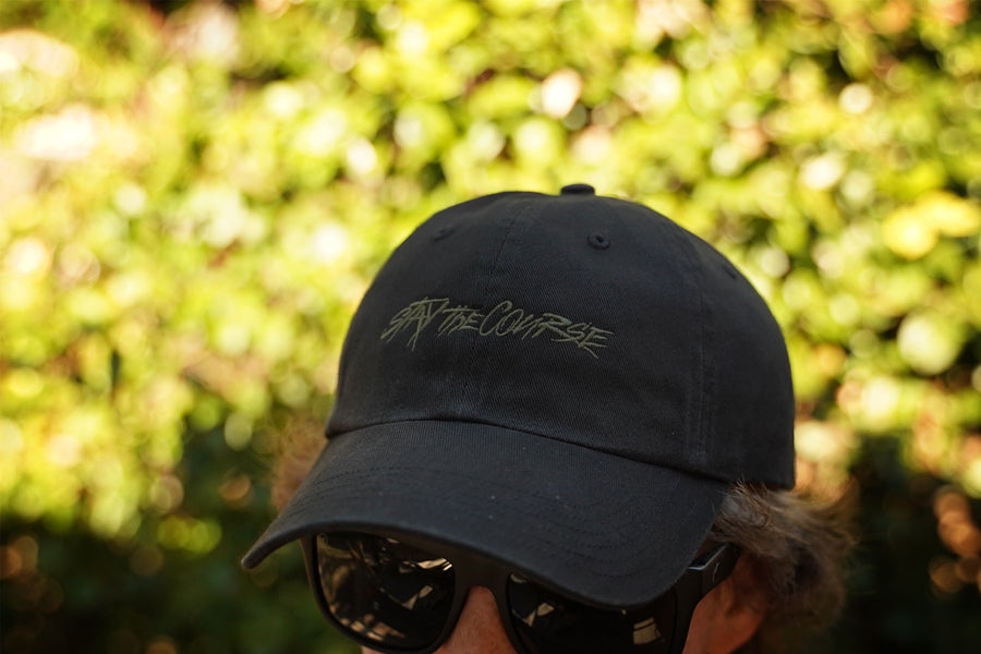 Person wearing a black cap with text and sunglasses, against a blurred green background