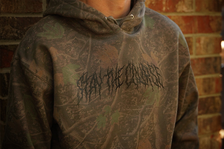 Darkwood tree camouflage hoodie with text design worn by a person, against a brick wall background.
