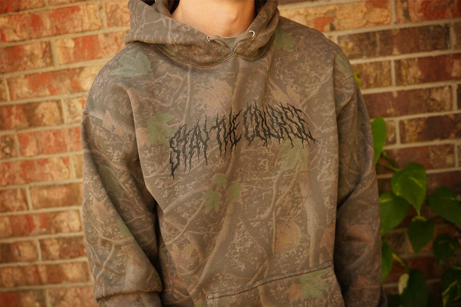 Camouflage hoodie with text design worn by a person against a brick wall.