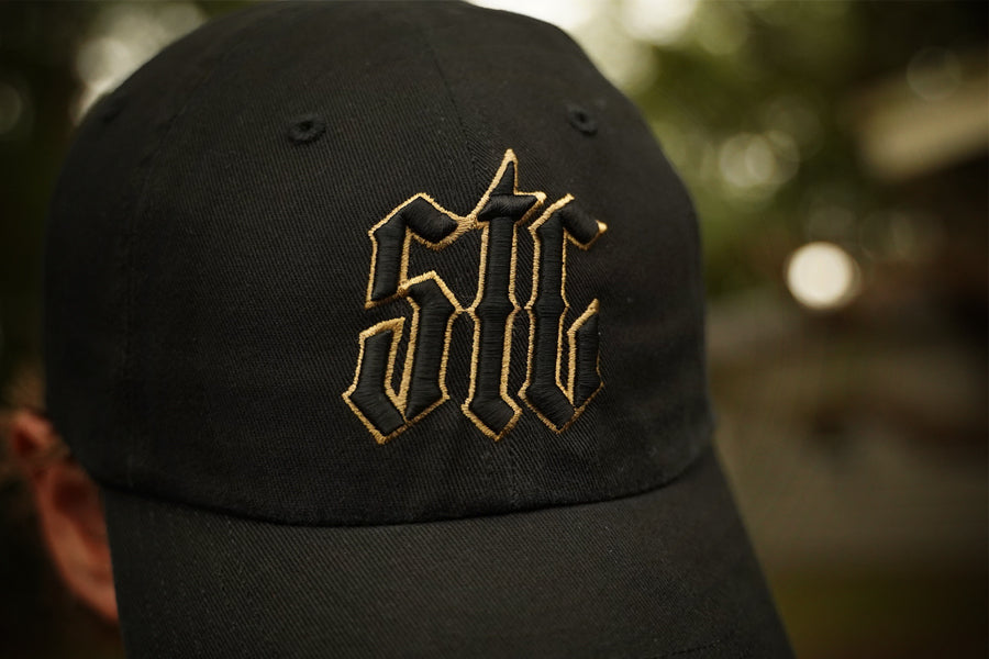Black cap with gold embroidered logo against a blurred natural background