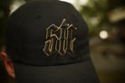 Black cap with gold embroidered logo against a blurred natural background