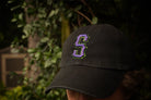 Person wearing a black cap with a purple 'S' logo against a blurred natural background