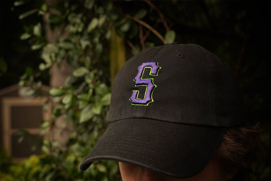 Person wearing a black cap with a purple 'S' logo against a blurred natural background