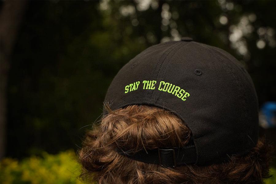 Black cap with 'Stay the Course' text worn by a person outdoors