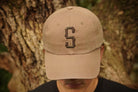 Beige cap with a letter 'S' worn by a person against a tree background