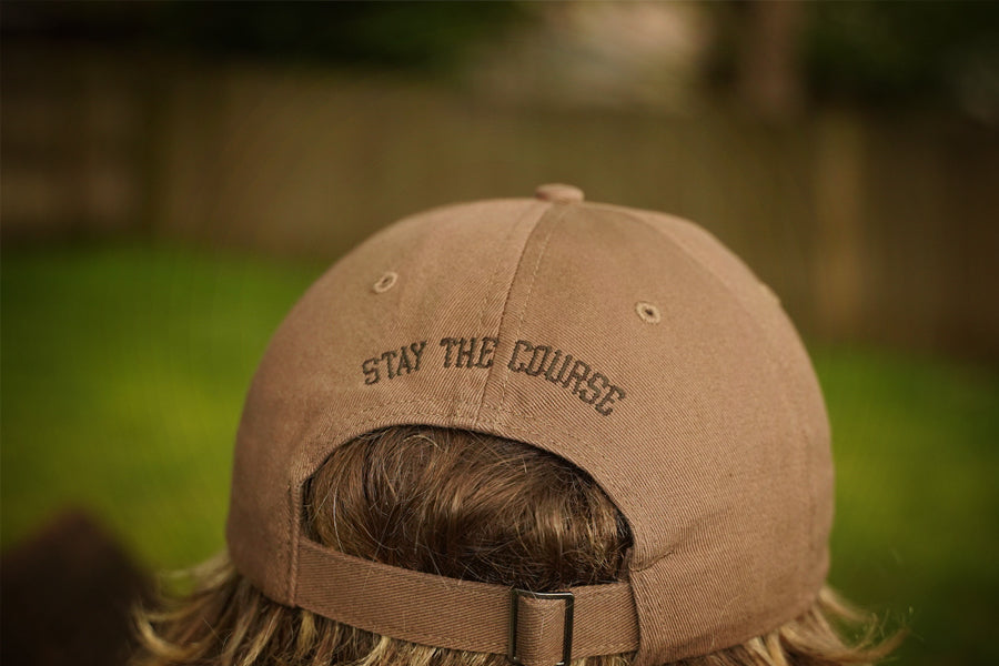 Brown cap with 'Stay the Course' text on a blurred outdoor background
