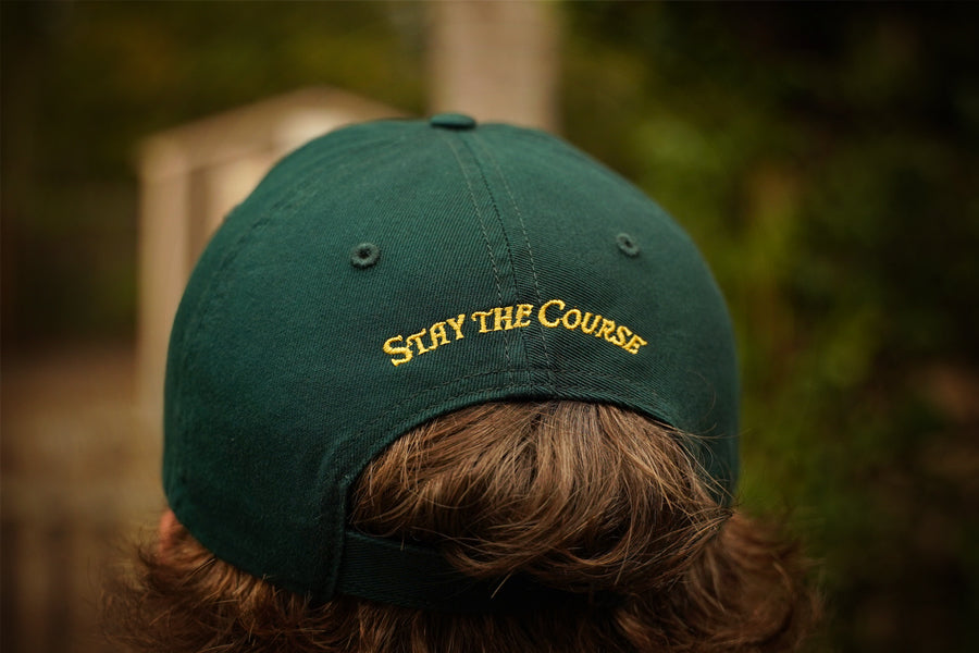 Green cap with 'Stay the Course' text worn by a person, blurred background