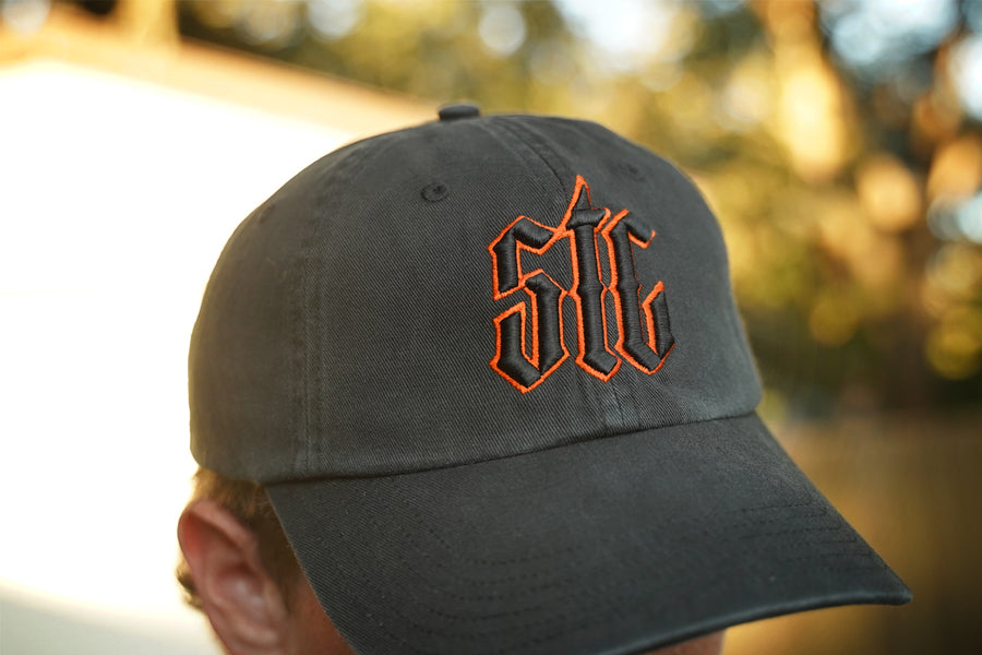 Person wearing a black hat with orange and black "stc" puff embroidery on the front, in an outdoor setting