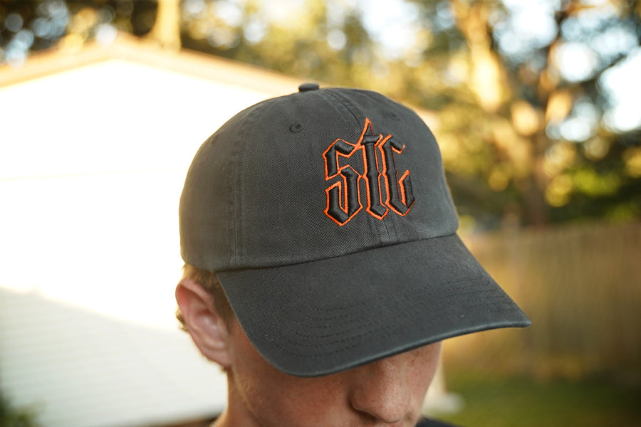 Person wearing a black hat with orange and black "stc" puff embroidery on the front, in an outdoor setting