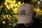 Beige cap with 'STG' logo worn by a person against a blurred natural background
