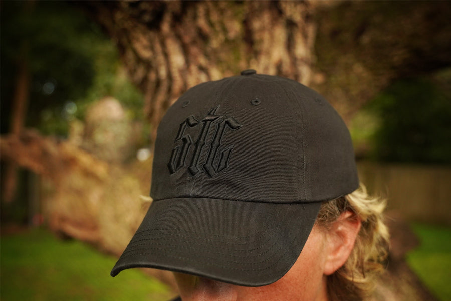 Person wearing a black cap with a logo, standing outdoors with a blurred background