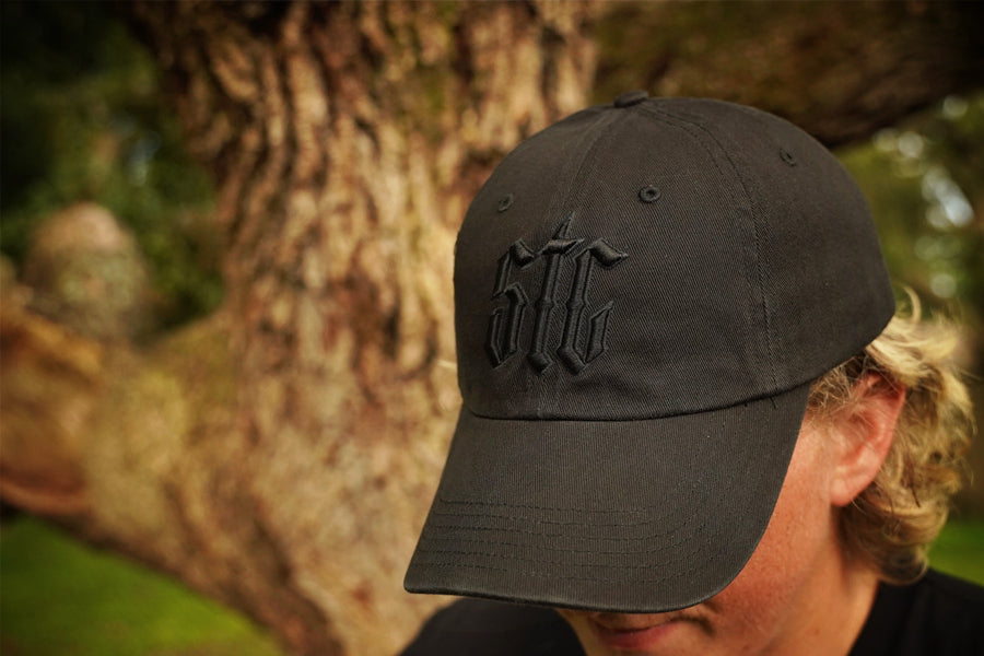 Person wearing a black cap with a logo, standing against a tree.