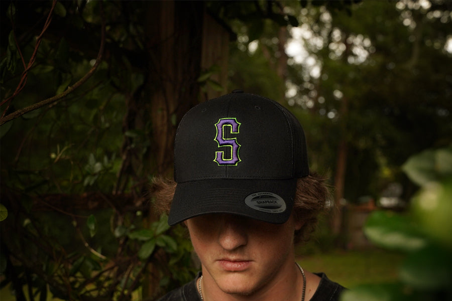 Person wearing a black cap with a 'S' logo in a forest setting