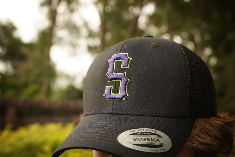 Black trucker cap with a purple and green logo worn by a person outdoors.