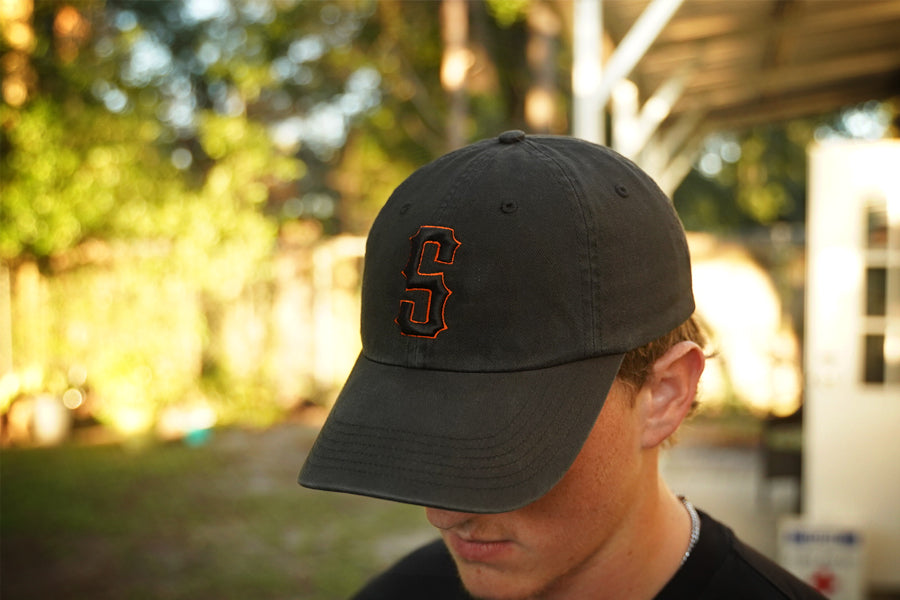 Person wearing a black hat with orange and black "s" puff embroidery on the front, in an outdoor setting