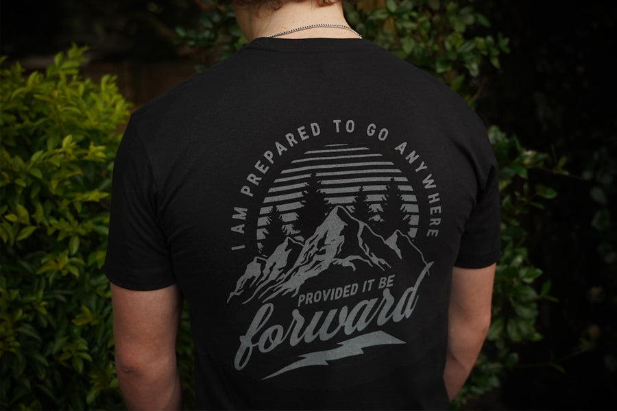 Person wearing a black t-shirt with a mountain design and text on the back, standing against a green leafy background.