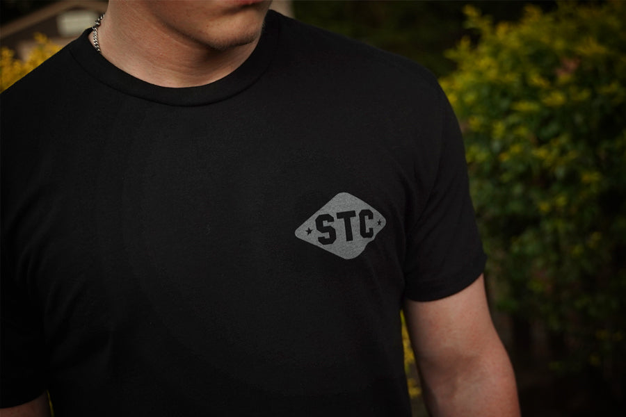 Person wearing a black t-shirt with 'STC' logo against a blurred green background