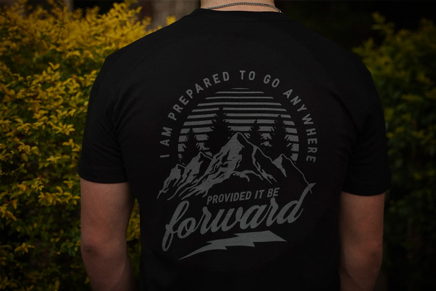 Person wearing a black t-shirt with a mountain design and text on the back, standing in front of greenery.