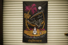 A black fabric banner featuring a colorful graphic of a skeleton reclining in a palm tree hammock on a tropical island, with the text 'staythecourse' at the bottom, hanging against a beige corrugated metal wall.
