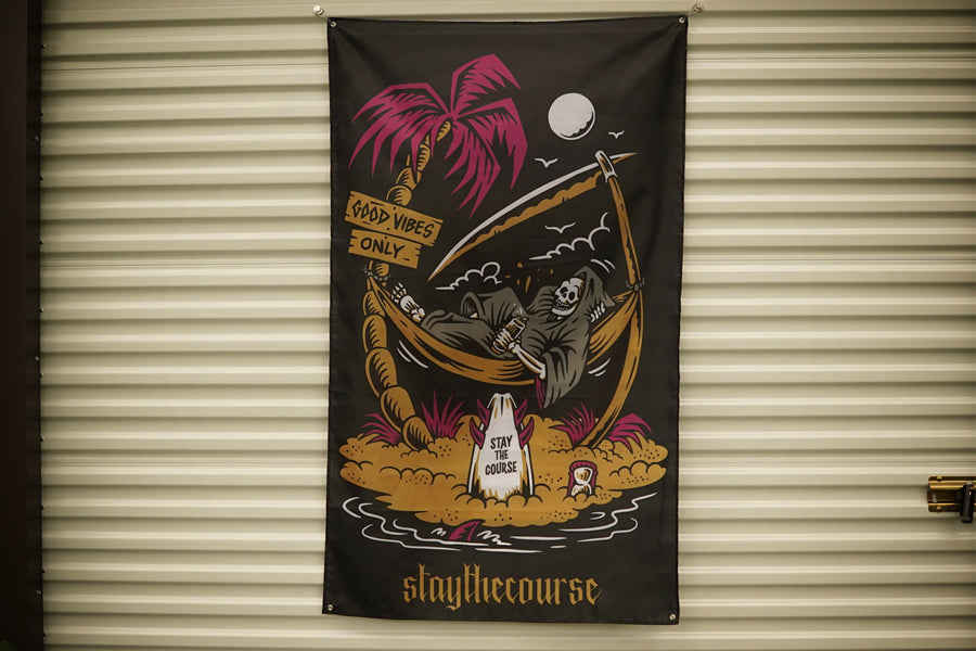 A black fabric banner featuring a colorful graphic of a skeleton reclining in a palm tree hammock on a tropical island, with the text 'staythecourse' at the bottom, hanging against a beige corrugated metal wall.