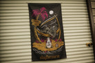 A black vertical banner featuring a grim reaper lounging in a palm tree hammock on a beach, with a pink palm tree, moon, and the text 'staythecourse' at the bottom, hanging against a beige corrugated metal wall.