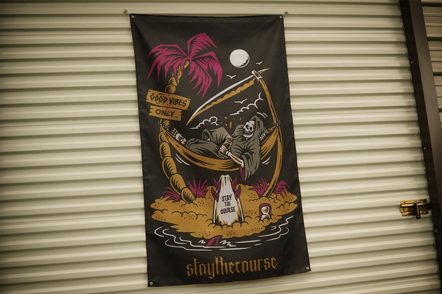 A black vertical banner featuring a grim reaper lounging in a palm tree hammock on a beach, with a pink palm tree, moon, and the text 'staythecourse' at the bottom, hanging against a beige corrugated metal wall.