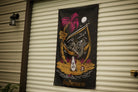 A vertical black banner hanging on a beige corrugated metal garage door. The banner features a colorful graphic illustration of a skeleton in a hammock under a palm tree, with the text 'slaythe course' at the bottom.
