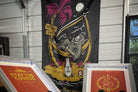 A large black fabric banner featuring a colorful graphic of a skeleton reclining in a hammock under a palm tree, displayed indoors in front of a white siding wall. Two framed prints are partially visible in the foreground.