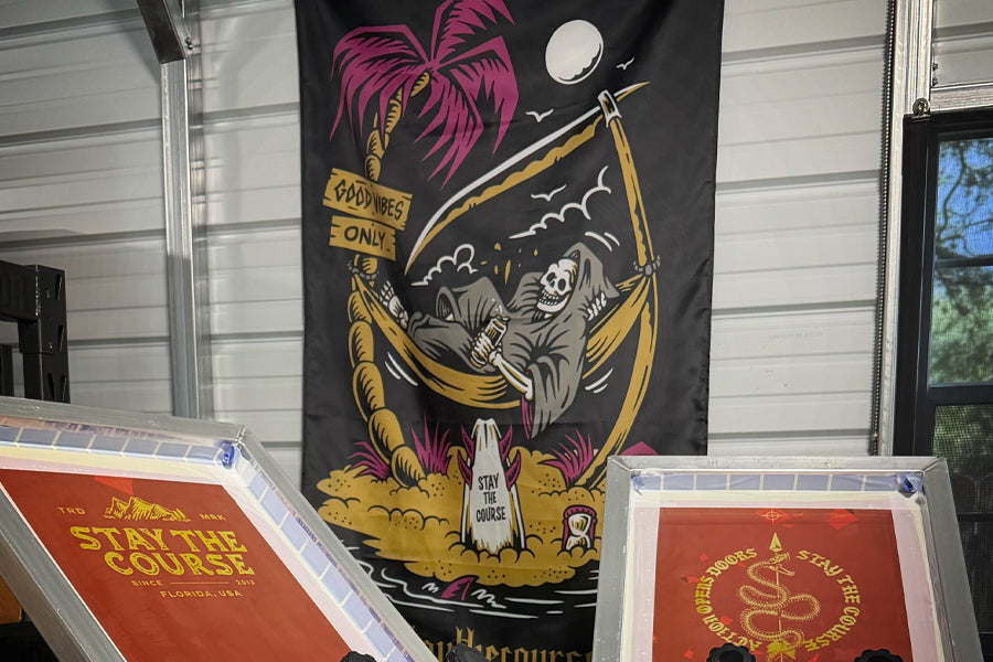 A large black fabric banner featuring a colorful graphic of a skeleton reclining in a hammock under a palm tree, displayed indoors in front of a white siding wall. Two framed prints are partially visible in the foreground.