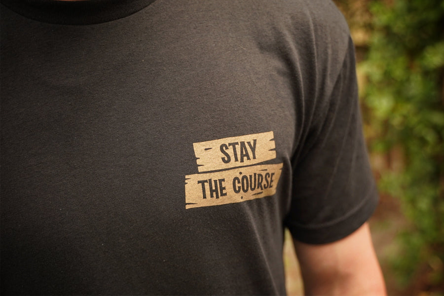 Black t-shirt with 'Stay The Course' text on a blurred natural background