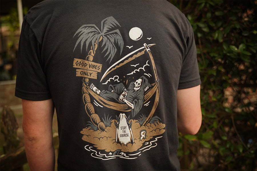 Person wearing a black t-shirt with a graphic design of a skeleton in a hammock, palm trees, and text.
