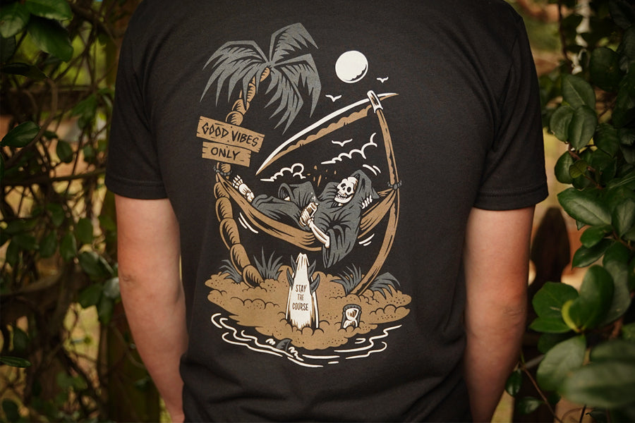 Person wearing a black t-shirt with a graphic design of a skeleton in a hammock, palm trees, and a moon.