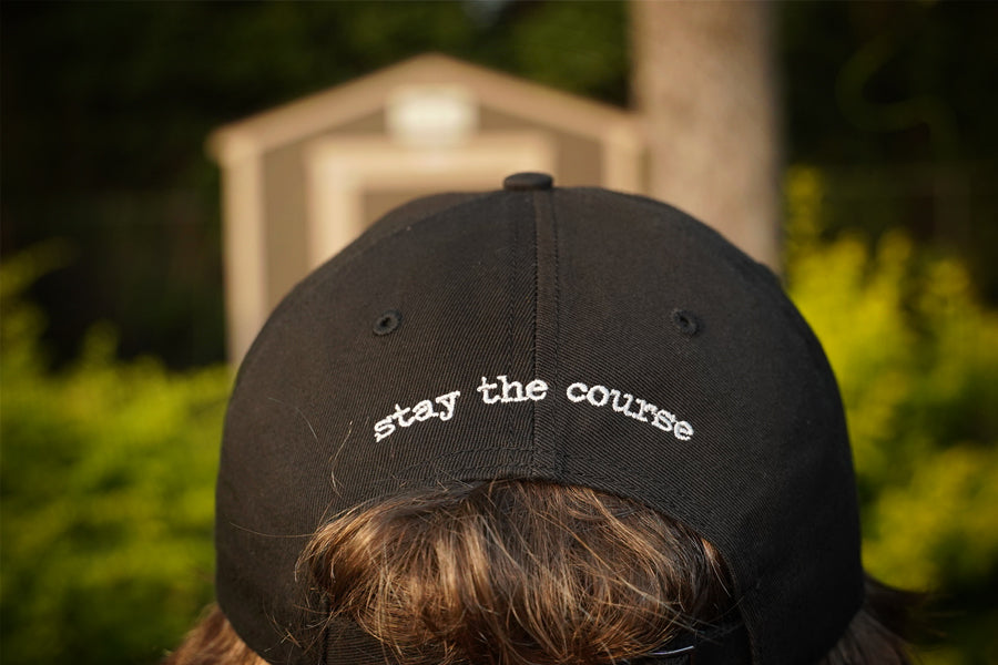 Black cap with 'stay the course' text worn by a person, with a blurred background of greenery and a building.