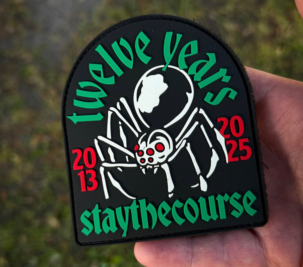 Patch with 'twelve years 2013-2025 staythecourse' text held by a hand against a blurred green background