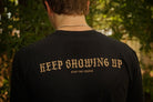 A person viewed from behind wearing a black t-shirt with the phrase 'KEEP SHOWING UP' and 'STAY THE COURSE' printed in a tan serif font, set against a blurred outdoor background of green foliage.