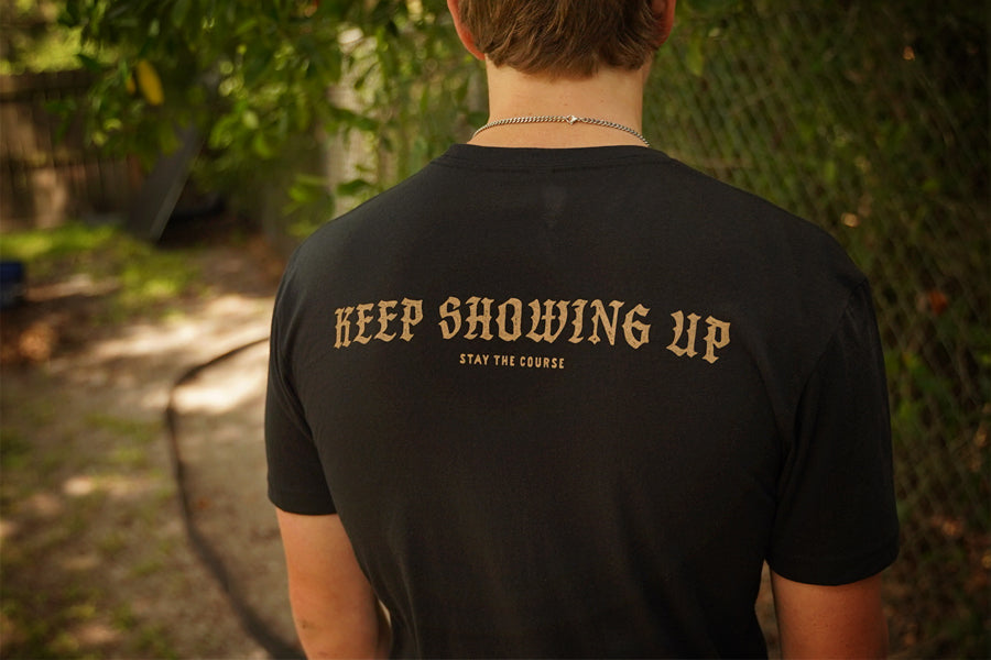 A person viewed from behind wearing a black t-shirt with 'KEEP SHOWING UP' and 'STAY THE COURSE' printed in gold on the back, set against a blurred outdoor background.