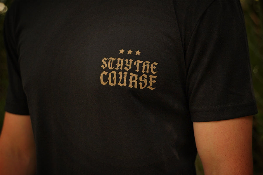 A close-up of a person wearing a black t-shirt with a gold printed graphic on the chest that reads 'STAY THE COURSE' with three stars above it.
