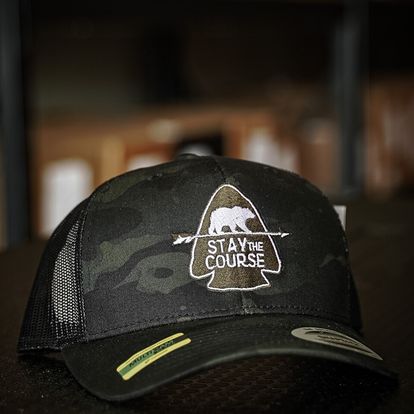 Black multicam trucker hat with 'Stay the Course' embroidered bear graphic on a blurred background