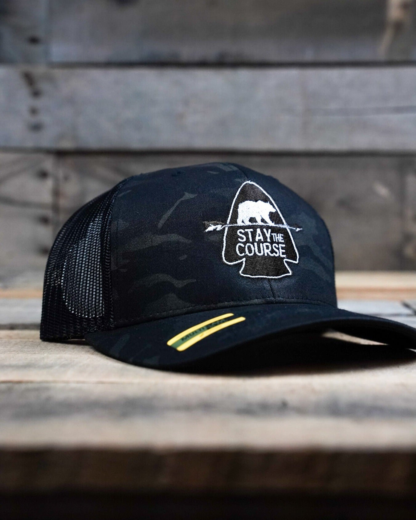 Black multicam cap with 'Stay the Course' logo on a wooden surface