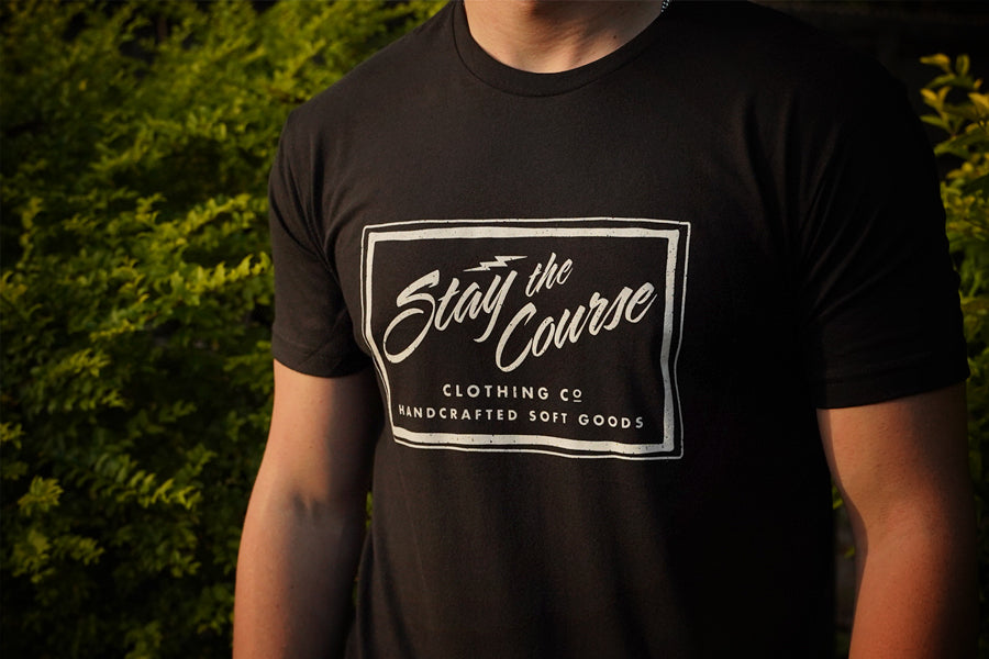 Person wearing a black t-shirt with 'Stay the Course' logo against a blurred green background