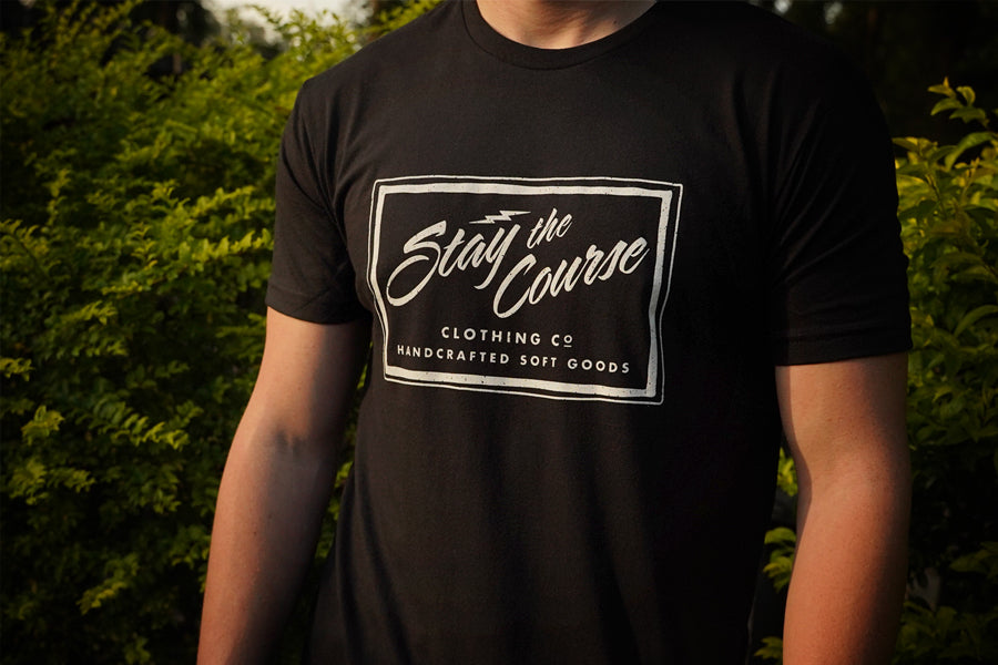 Person wearing a black t-shirt with a logo and text in an outdoor setting